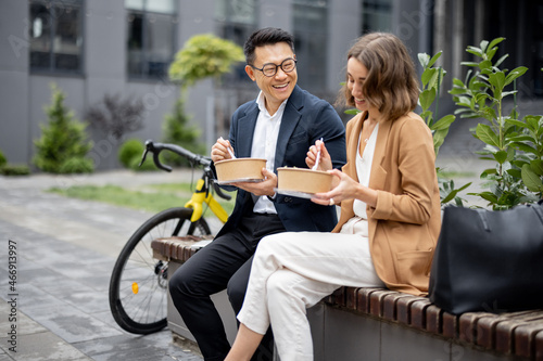 Asian businessman and caucasian businesswoman eating food and talking while having lunch at work. Concept of rest and break on job. Smiling business people sitting on bench in city