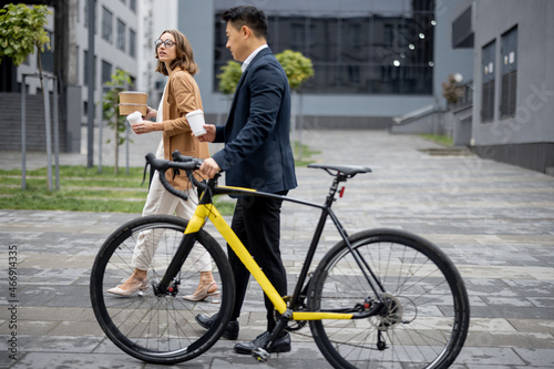 Asian businessman and caucasian businesswoman walking, drinking coffee and talking in city. Concept of business cooperation. Idea of freelance and remote work. Man with bicycle. Young woman with food