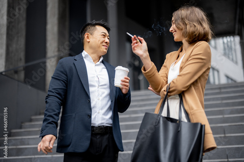 Asian businessman and smiling caucasian businesswoman smoking cigarettes during break at work on city street. Concept of modern successful business people. Idea of rest and break on job