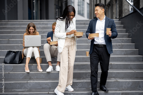 Businessman and businesswoman eating food on background of their working colleagues on city street. Concept of break on job. Idea of modern business people. Remote and freelance work