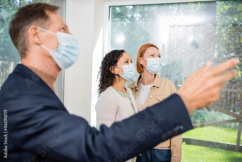 blurred realtor in medical mask pointing with hand near interracial lesbian couple inside of new house