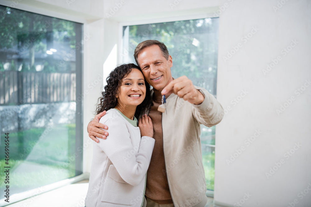 cheerful man holding blurred key and hugging african american wife in new house cheerful man holding blurred key and hugging african american wife in new house
