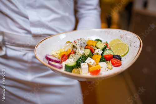 Fototapeta Naklejka Na Ścianę i Meble -  Waiter holds a plate with tasty dish, traditional Italian Greek salad. Waiter wearing white uniform in a restaurant.