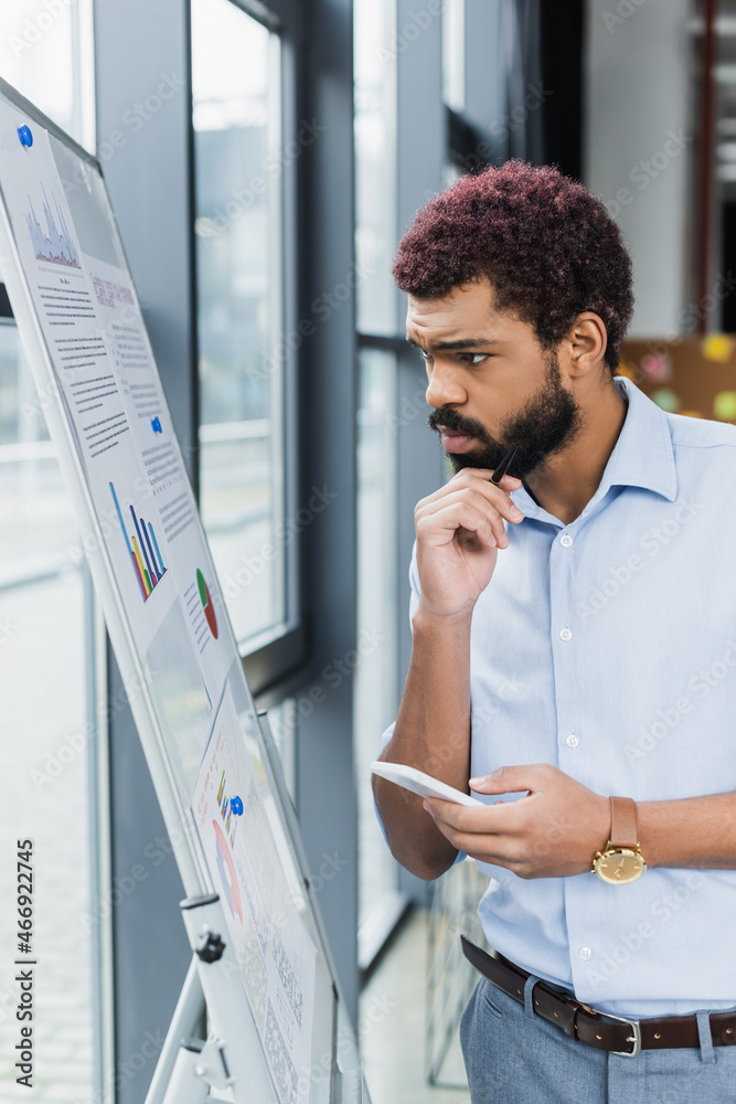 Pensive african american businessman holding smartphone and looking at flip chart in office