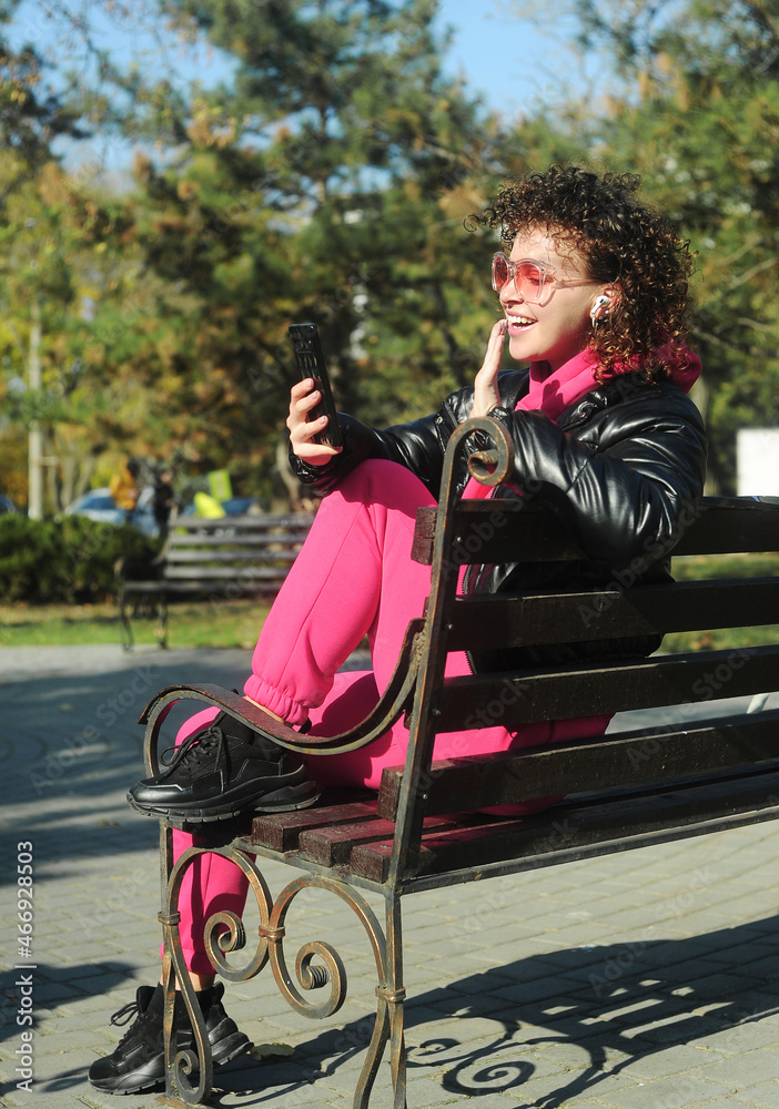 Woman holding mobile phone in hands and making selfie outdoors. Curly hair young woman in magenta sporty wear outside Woman holding mobile phone in hands and making selfie outdoors. Curly hair young woman in magenta sporty wear outside