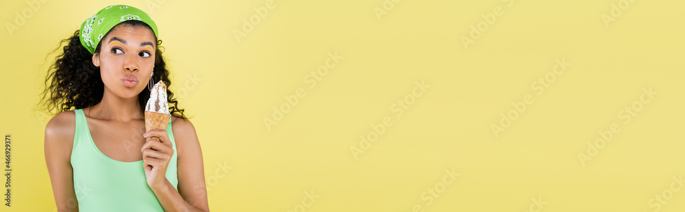 curly african american woman in green kerchief holding ice cream and pouting lips isolated on yellow, banner curly african american woman in green kerchief holding ice cream and pouting lips isolated on yellow, banner