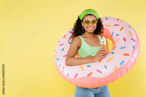 positive african american young woman standing with inflatable ring and ice cream cone isolated on yellow