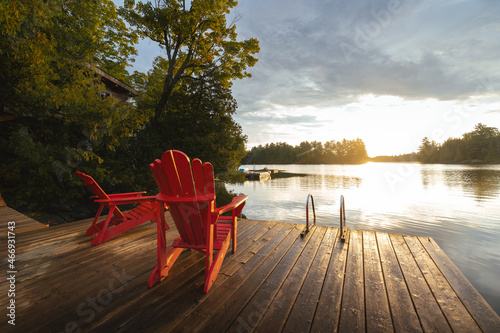 Two red chairs sit facing the sunrise on a dock photo