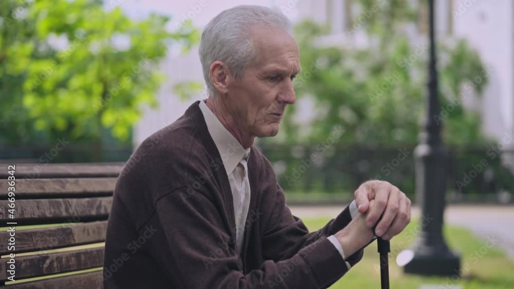 Lonely elderly male with walking stick sitting on park bench, feeling depression