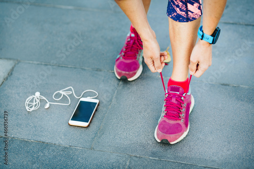 Runner woman tying running shoes laces getting ready for race on run track with smartphone and earphones for music listening on mobile phone. Athlete preparing for cardio training. Feet on ground