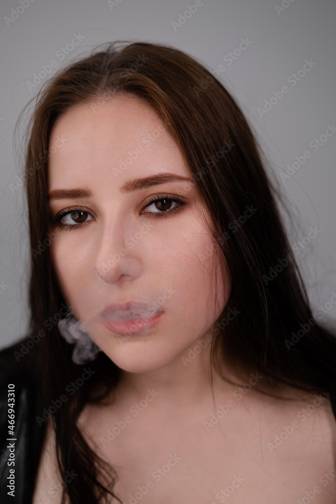 young brunette woman in black leather jacket exhales white smoke. beautiful girl millenial on grey background smoking. bad habit