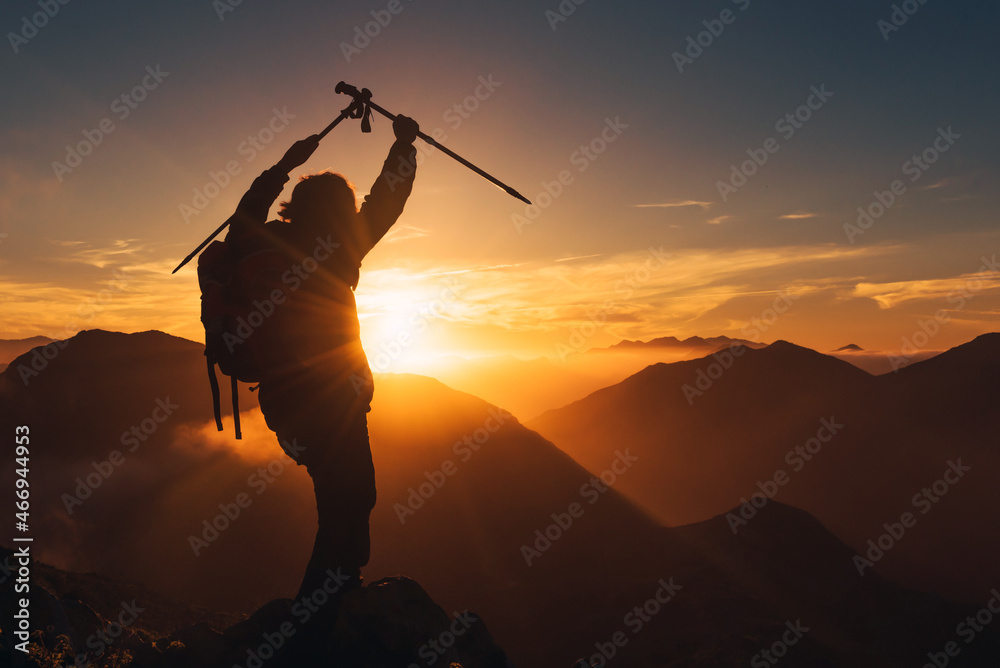 backlit silhouette of hiker with backpack raising his arms in victory sign while watching the sunset. mountain sport concept. backlit silhouette of hiker with backpack raising his arms in victory sign while watching the sunset. mountain sport concept.