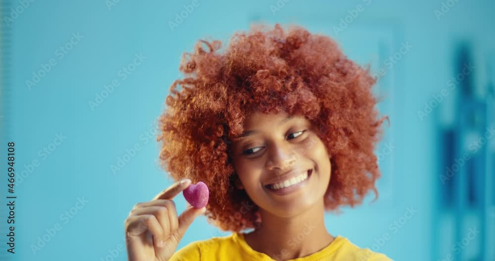 Slow motion close up of carefree African American female model with curly hair holding sweet candy and smiling at camera in studio. Joyful young black woman with brown afro hairstyle showing candy Slow motion close up of carefree African American female model with curly hair holding sweet candy and smiling at camera in studio. Joyful young black woman with brown afro hairstyle showing candy
