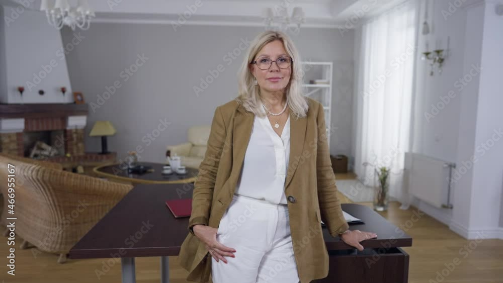 Blond Caucasian woman in eyeglasses standing in office at table looking at camera smiling. Portrait of confident professional psychologist posing indoors. Lifestyle and confidence concept Blond Caucasian woman in eyeglasses standing in office at table looking at camera smiling. Portrait of confident professional psychologist posing indoors. Lifestyle and confidence concept