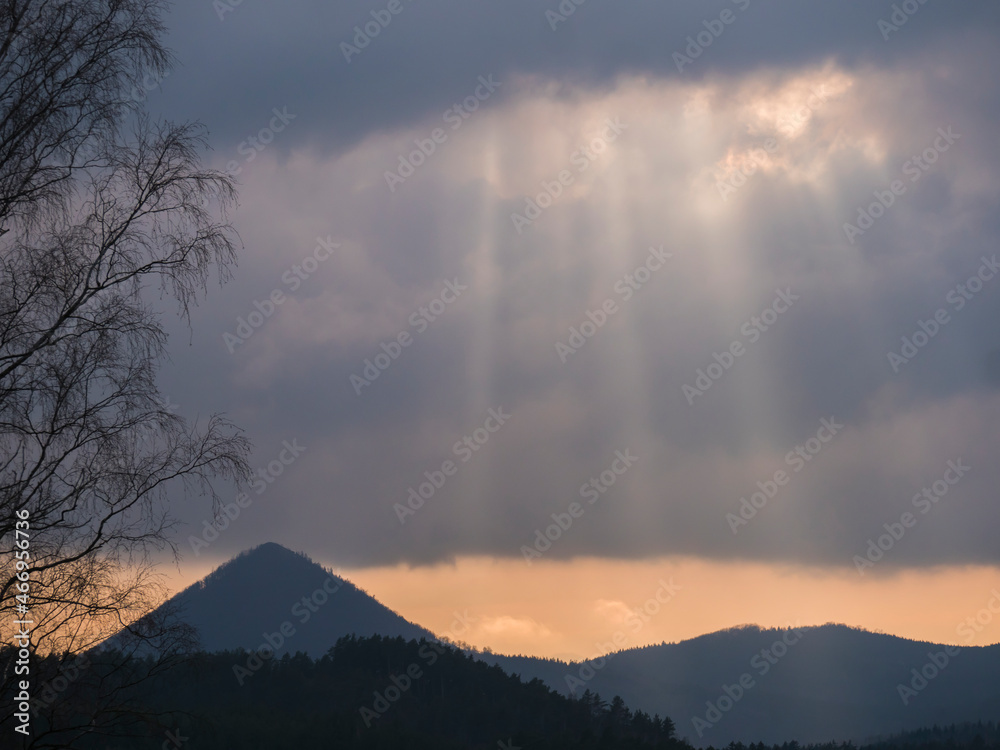 Overcast sky before sunset with sun rays at forest landscape with hill Klic, Kleis. Sunlight shining through hole of clouds to dark scene of mountain range. Moody natural background, copy space Overcast sky before sunset with sun rays at forest landscape with hill Klic, Kleis. Sunlight shining through hole of clouds to dark scene of mountain range. Moody natural background, copy space