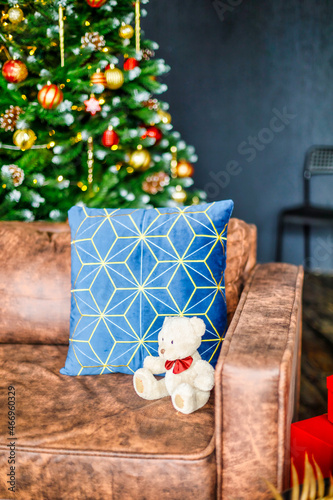 The room is decorated with New Year's decor. A festive tree decorated with New Year's toys next to a brown sofa. On the couch, a blue pillow and a white teddy bear