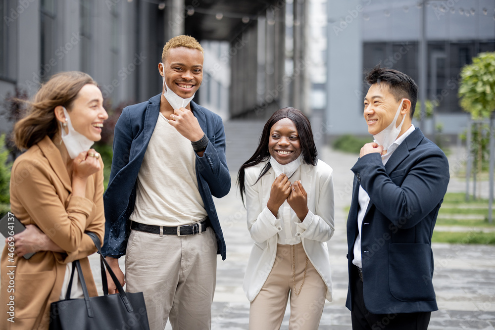 Multiracial business people taking off medical masks and talking on city street. Concept of health protection during Coronavirus pandemic. Joyful businessmen and businesswomen wearing formal wear Multiracial business people taking off medical masks and talking on city street. Concept of health protection during Coronavirus pandemic. Joyful businessmen and businesswomen wearing formal wear