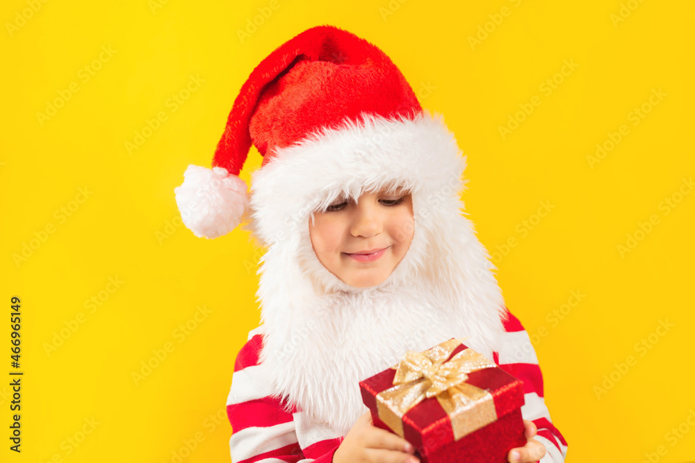 Funny child toddler in a Santa hat with red gift , yellow background. Holidays, Traditions, Christmas concept. Funny child toddler in a Santa hat with red gift , yellow background. Holidays, Traditions, Christmas concept.