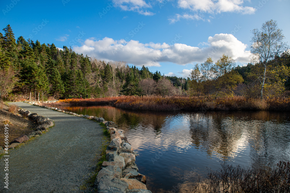 A walking trail of crushed stone and gravel along a wide river. The trail has water on one side and a rock face cliff on the other side. The stream is calm and reflects the mountain covered in trees A walking trail of crushed stone and gravel along a wide river. The trail has water on one side and a rock face cliff on the other side. The stream is calm and reflects the mountain covered in trees