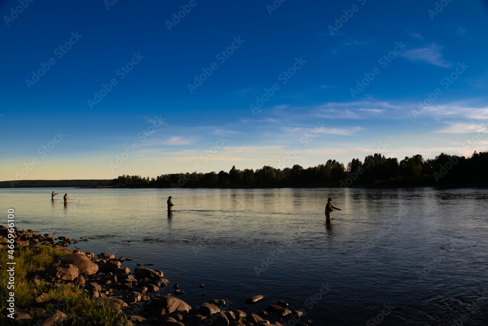 Male salmon anglers standing in a river up to their waist with long fishing rods wearing waders, fly hats, and vests. The evening sky is blue and pink with smooth river water. 