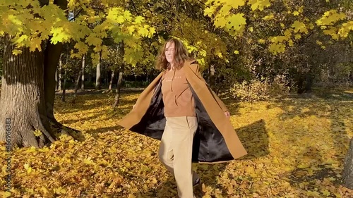 A woman in a coat dances and spins, arms outstretched in the sunny autumn forest
