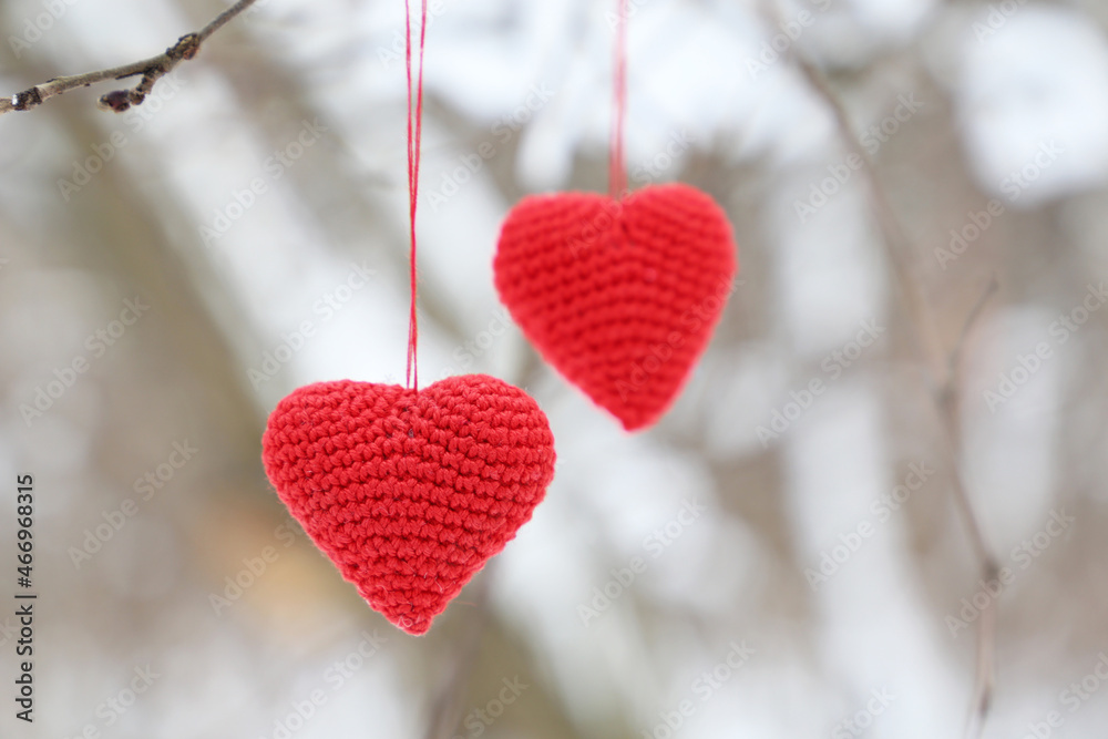 Fototapeta premium Love hearts hanging on a tree branch in winter forest. Two red knitted Valentine hearts, symbol of romantic love, background for holiday