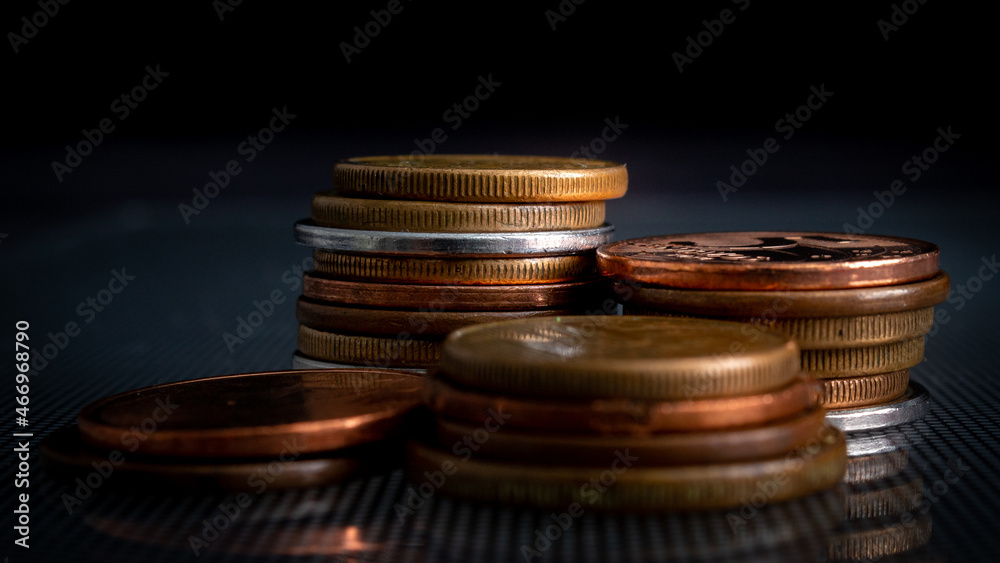 VARIAS MOEDAS EMPINHADAS EMCIMA DE UM VIDRO, COM O FUNDO ESCURO, COM FOCO EM APENAS ALGUMAS MOEDAS.