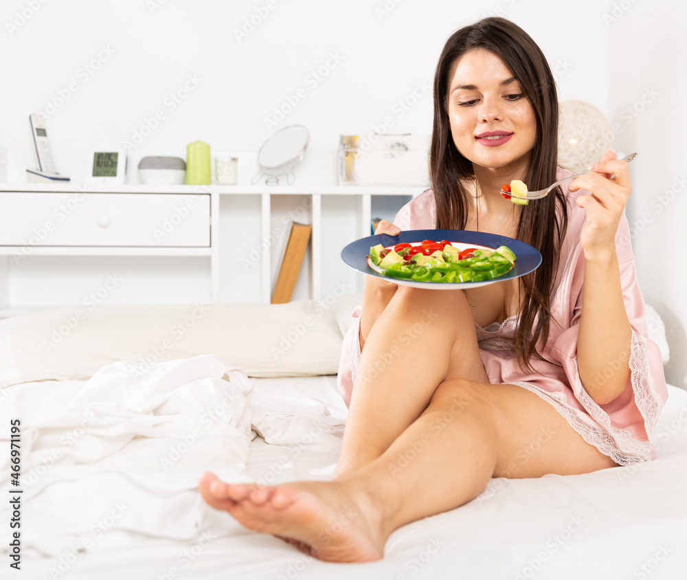 Portrait of lady in bathrobe eating vegetable salad in bed at home alone