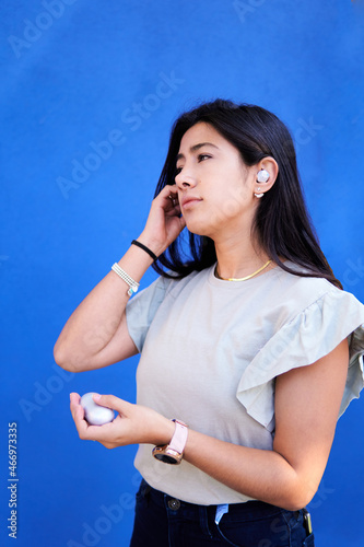 happy young woman, slim, putting on wireless headphones, on blue background with copy space, technology, portrait photo