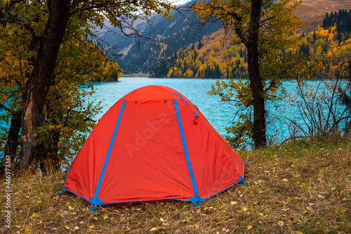 Red tent near beautiful mountains lake with turquoise water and colorful forest around. Tourism, trekking in autumn season concept. Camp in mountains. Zhasylkol lake in Dzungarian Alatau, Kazakhstan.