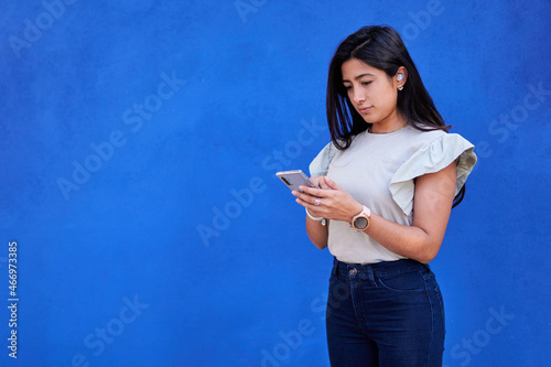 young latina female delegate with brown skin, looking at cell phone and typing, with wireless headset, on a blue background, copy space, technology