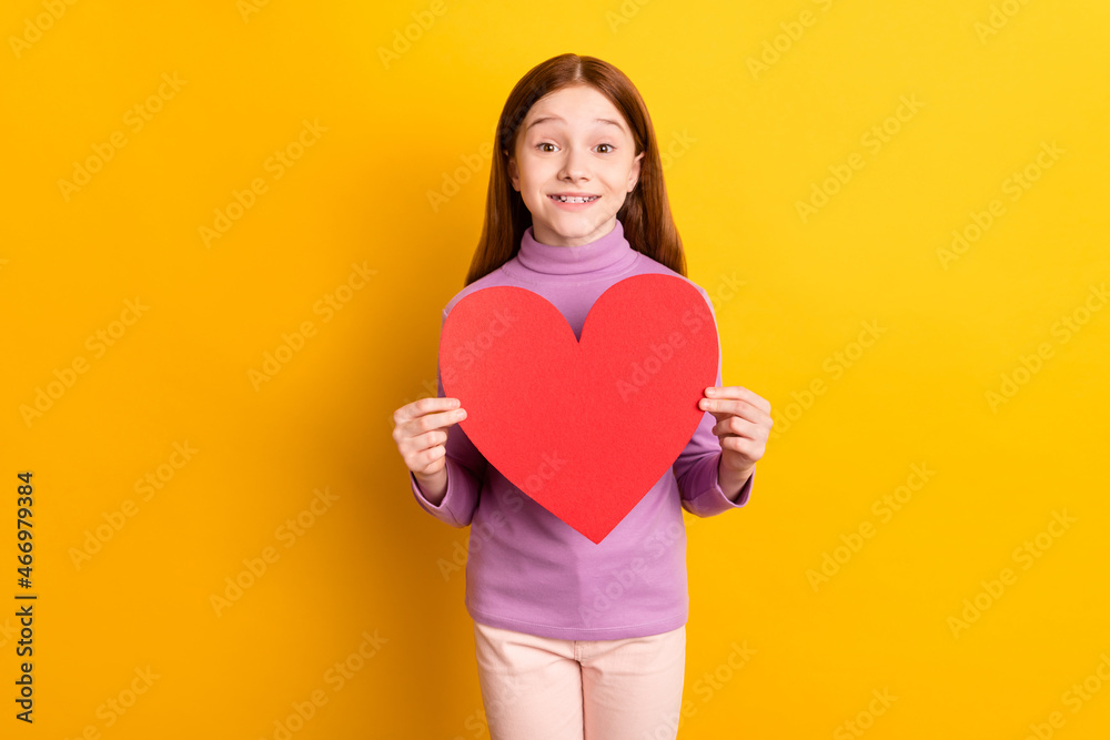 Portrait of attractive cheerful red-haired girl holding in hands big large heart card symbol isolated over bright yellow color background Portrait of attractive cheerful red-haired girl holding in hands big large heart card symbol isolated over bright yellow color background