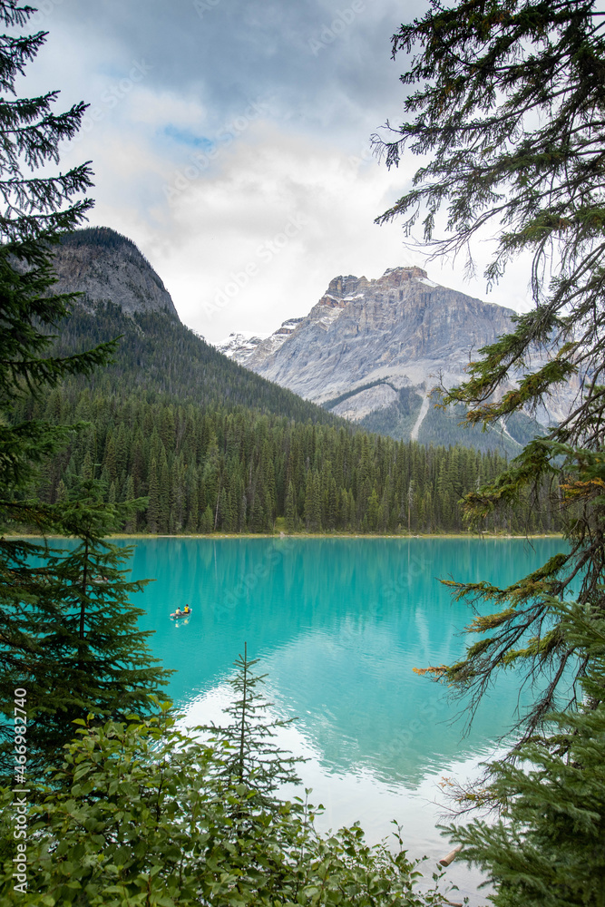 Photo of Emerald lake located in Yoho National Park. There are pine trees in the foreground. Beyond the trees there is emerald lake, a couple who are in a canoe and mountains in the background. 