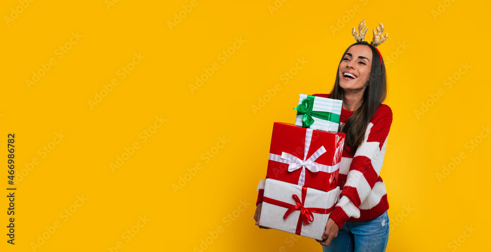 Cute smiling excited young brunette woman in deer hat, with many colorful Christmas gift boxes in hands isolated on yellow background. - obrazy, fototapety, plakaty 