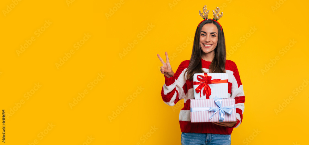 Cute smiling excited young brunette woman in deer hat, with many colorful Christmas gift boxes in hand is showing victory sign isolated on yellow background. Cute smiling excited young brunette woman in deer hat, with many colorful Christmas gift boxes in hand is showing victory sign isolated on yellow background.
