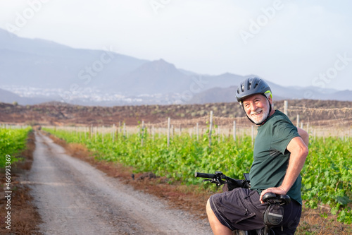 Smiling senior man enjoying healtlhy lifestyle riding in a vineyard at sunset light with his bicycle. Elderly bearded man wearing sport helmet in countryside, mountains in background photo