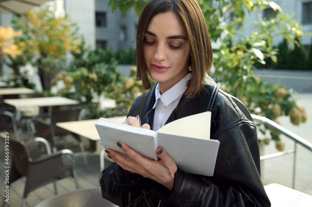 Confident portrait of focused young woman writing on white notepad while sitting outdoors on urban public background Confident portrait of focused young woman writing on white notepad while sitting outdoors on urban public background