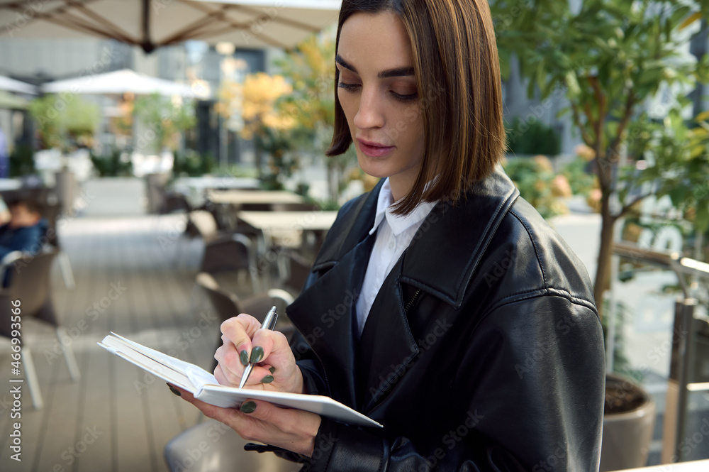 Confident portrait of concentrated young business woman, copywriter, journalist, writing on white notepad while sitting outdoors on urban public background. Close-up Confident portrait of concentrated young business woman, copywriter, journalist, writing on white notepad while sitting outdoors on urban public background. Close-up
