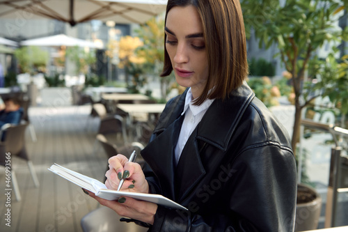 Confident portrait of concentrated young business woman, copywriter, journalist, writing on white notepad while sitting outdoors on urban public background. Close-up © Taras Grebinets