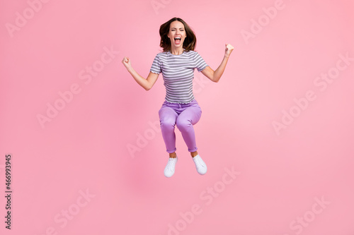 Photo of funny lucky young woman dressed striped clothes jumping high rising fists smiling isolated pink color background