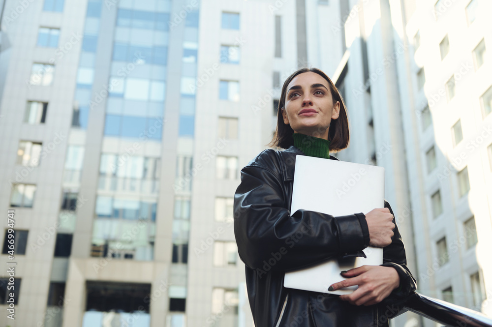 Lifestyle portrait of successful self-confident motivated young business person, attractive freelance woman holding laptop, looking up posing against modern corporate high-rise buildings background Lifestyle portrait of successful self-confident motivated young business person, attractive freelance woman holding laptop, looking up posing against modern corporate high-rise buildings background