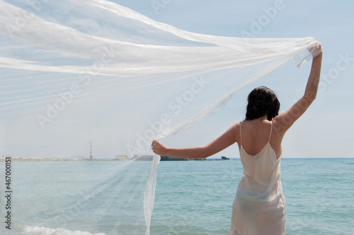 Rear view of a woman standing on a beach holding a sheer veil over her head