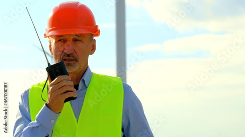 Wind turbine produces energy at offshore station. Experienced technician in orange helmet uses radio set walking against rotating propeller photo