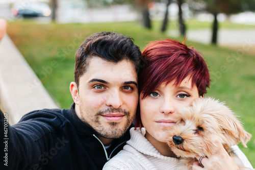 A young, handsome, light-eyed couple taking a selfie outdoors with their dog while walking in the city park.