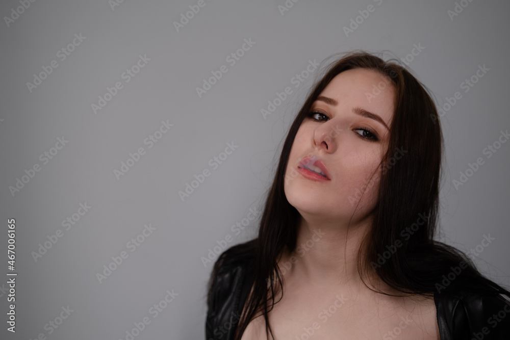 young brunette woman in black leather jacket exhales white smoke. beautiful girl millenial on grey background smoking. bad habit young brunette woman in black leather jacket exhales white smoke. beautiful girl millenial on grey background smoking. bad habit