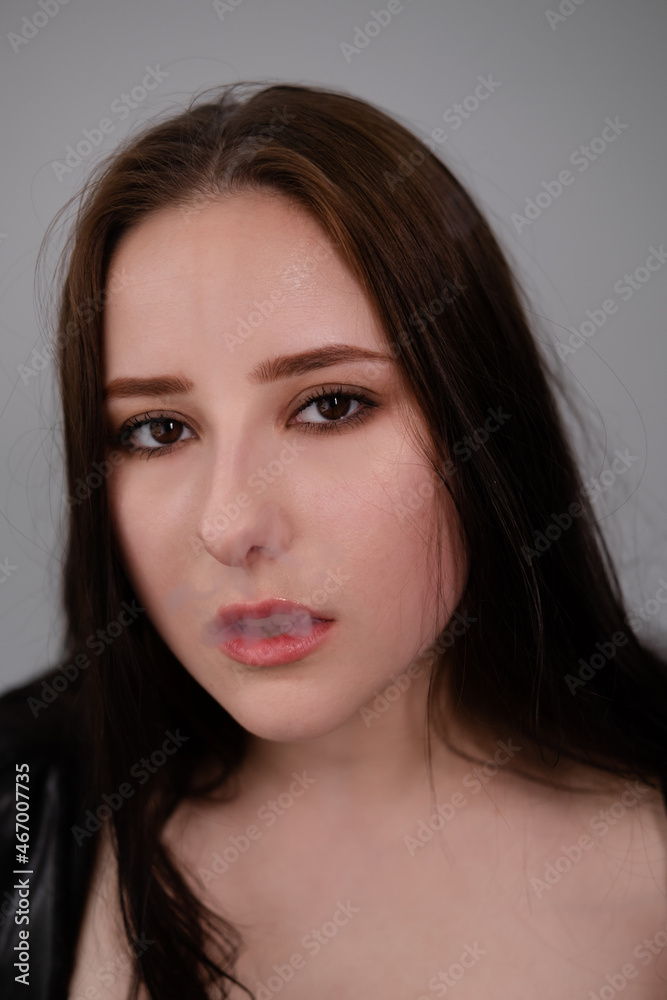 young brunette woman in black leather jacket exhales white smoke. beautiful girl millenial on grey background smoking. bad habit young brunette woman in black leather jacket exhales white smoke. beautiful girl millenial on grey background smoking. bad habit