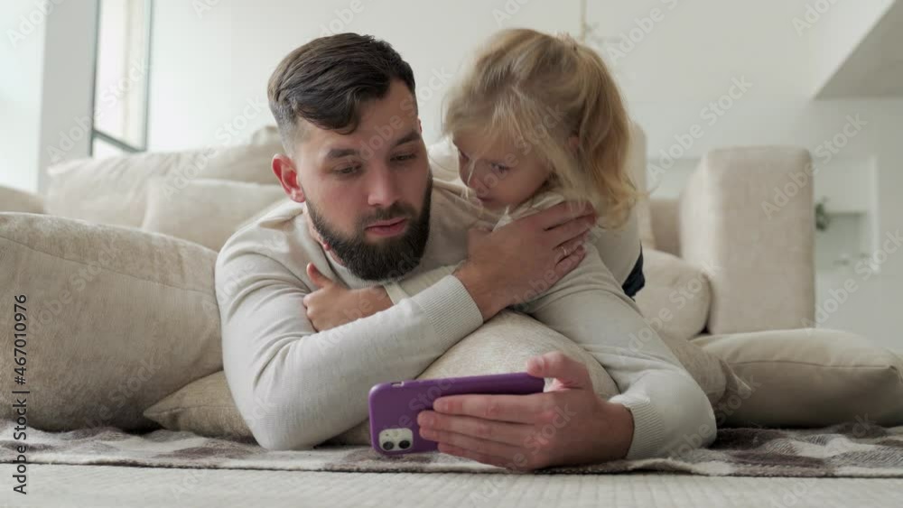 Bearded cheerful dad and his little daughter watching videos on a smartphone and laughing merrily lying on the floor in the living room Bearded cheerful dad and his little daughter watching videos on a smartphone and laughing merrily lying on the floor in the living room