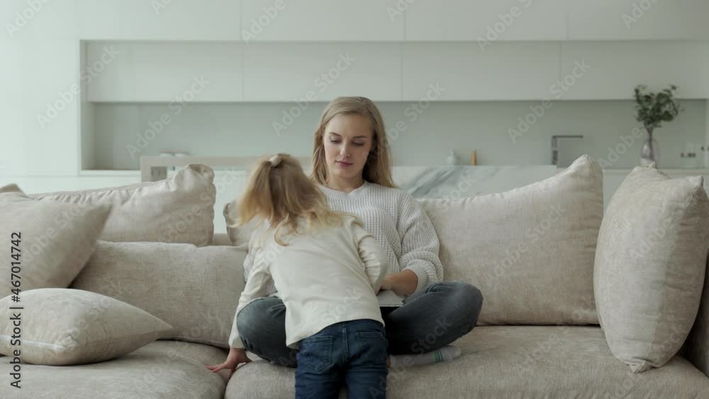 Female entrepreneur focused on working on a laptop on the couch while her daughter plays and prevents her mom from working Female entrepreneur focused on working on a laptop on the couch while her daughter plays and prevents her mom from working