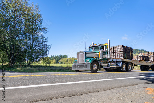 Classic compact powerful old style green big rig semi truck transporting fastened cargo on flat bed semi trailer running on the road at sunshine