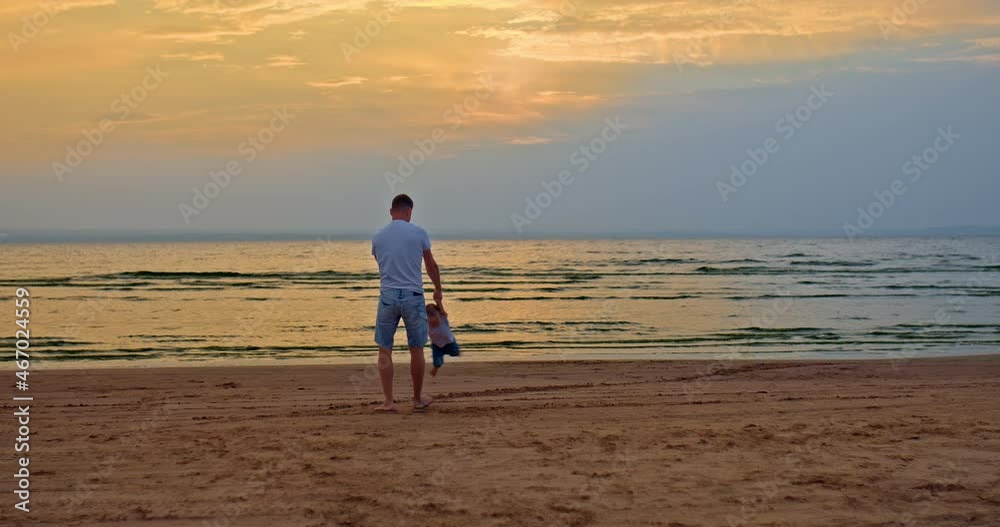 Caring father plays with his child, circling his son by the hands. Dad and little son are playing on the seashore, walking on the sand with their bare feet. Dad and son are walking at sunset. Happy Caring father plays with his child, circling his son by the hands. Dad and little son are playing on the seashore, walking on the sand with their bare feet. Dad and son are walking at sunset. Happy